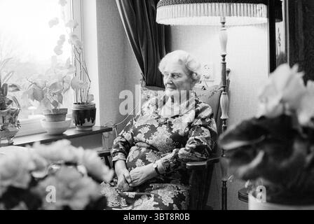 Portrait de femme 80+ assise dans le salon, Whizgle News, Dutch Desk, pays-Bas, 1950 - 2000 sur 08-12-1989. Ce sont les éléments de l'image. Dans un cadre intérieur confortable, une femme âgée s'assoit gracieusement dans un fauteuil en bois, dégageant une présence calme. Elle est habillée d'une robe à motifs floraux qui met en valeur des tons doux de rose et de bleu, s'harmonisant avec l'ambiance chaleureuse et accueillante de la pièce. Ses cheveux sont coiffés dans des boucles douces, et elle a une expression douce et sereine sur son visage, suggérant sagesse et expérience. Autour d'elle sont des plantes vertes luxuriantes, avec quelques fleurs en pot qui ajoutent spl Banque D'Images