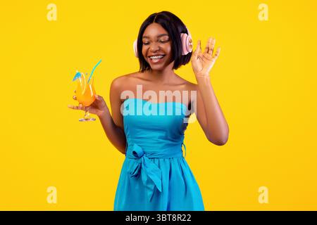Femme noire dans un ensemble élégant tenant un cocktail exotique et appréciant musique préférée dans les écouteurs sur fond orange Banque D'Images