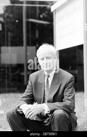 Portrait d'un homme près d'une pancarte avec inscription (Moret Ernst & Young), Whizgle News, Dutch Desk, pays-Bas, 1950 - 2000 on 09-1995. L'image montre ces rubriques. Dans la scène, un homme est assis confortablement sur le sol dans un espace extérieur bien entretenu, caractérisé par une pelouse verte luxuriante. Il est habillé formellement dans un costume sombre avec une chemise blanche éclatante et une cravate à motifs qui ajoute un contraste subtil à sa tenue. Ses cheveux sont gris, soigneusement peignés, et il a une expression calme, suggérant un sentiment d'équilibre et de professionnalisme. L'arrière-plan présente un bâtiment moderne, avec un verre clair et réfléchissant su Banque D'Images