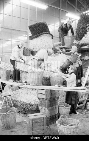 Chanson de groupe et amitié avec flotteur de parade Flower exposition de fleurs de parade, Whizgle News, Dutch Desk, pays-Bas, 1950 - 2000 le 21-04-1982. L'image inclut ces rubriques. La scène se déroule dans un espace intérieur spacieux et bien éclairé, mis en valeur par de grandes fenêtres permettant à la lumière naturelle de filtrer, créant une atmosphère chaleureuse et accueillante. L'objectif principal est un affichage floral imposant, prenant la forme d'un bonhomme de neige fantaisiste orné d'un grand chapeau haut de gamme. Le bonhomme de neige est méticuleusement construit à partir de matériaux floraux blancs et verts, qui ajoutent texture et dimension. Dans cet environnement animé Banque D'Images
