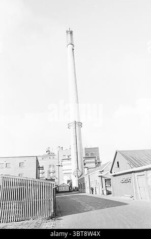 Cheminée en papier Gelder, Whizgle News, Dutch Desk, pays-Bas, 1950 - 2000 sur 15-02-1965. Ce sont les éléments de l'image. La scène présente un paysage industriel saisissant caractérisé par une cheminée cylindrique haute qui s'élève bien en évidence contre un ciel pâle et nuageux. Sa surface est élégante et métallique, réfléchissant subtilement la lumière et dominant les bâtiments environnants. La cheminée est placée sur une toile de fond d'une grande structure industrielle rectangulaire qui a un design quelque peu utilitaire, avec divers tuyaux et évents qui suggèrent une installation opérationnelle. Po Banque D'Images