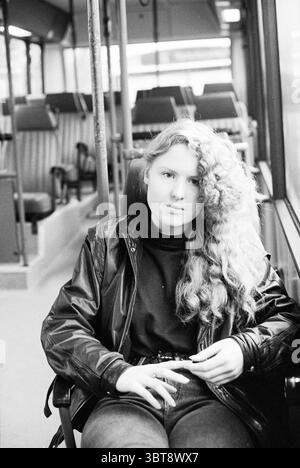 Olga Racwitz dans le bus, Whizgle News, Dutch Desk, pays-Bas, 1950 - 2000 le 22-12-1992. Voici les rubriques de l'image. Dans un intérieur un peu faiblement éclairé, une jeune femme est assise dans un bus, créant un point focal frappant dans la scène tamisée. Ses longs cheveux bouclés descendent en cascade d'un côté de son visage, étincelant doucement sous la lumière disponible. Elle porte une veste en cuir noir sur un haut sombre, suggérant un comportement élégant mais décontracté. Les sièges d'autobus, disposés en rangées soignées, sont d'une teinte brune discrète, avec une deuxième teinte plus foncée sur les coussins supérieurs. L'intérieur du bus dispose d'un design simple, avec p Banque D'Images