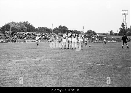 Football Haarlem pays-Bas, Whizgle News, Dutch Desk, pays-Bas, 1950 - 2000 en 1964. Ces rubriques apparaissent dans l'image. La scène capture un moment dynamique sur un terrain de sport, où un groupe de joueurs est aligné dans une position défensive. Vêtus de maillots rayés, les joueurs affichent des expressions ciblées alors qu'ils se préparent à un coup de pied imminent. Leurs uniformes contrastent avec le vert luxuriant de l'herbe sous eux, qui est légèrement usé, indiquant un match bien joué. En arrière-plan, une foule modeste est visible, avec de petits groupes de spectateurs assis dans des gradins ou debout près Banque D'Images