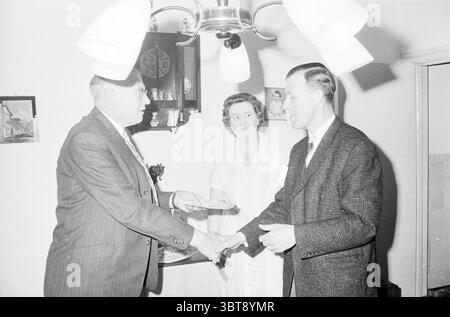Cérémonie de remise des prix de la carte de sécurité, Whizgle News, Dutch Desk, pays-Bas, 1950 - 2000 le 22-06-1961. L'image inclut ces rubriques. Dans un cadre intérieur chaleureusement éclairé, trois individus sont engagés dans un moment d'interaction, souligné par un sens de formalité ou de célébration. L'espace a une sensation confortable et accueillante, renforcée par un fond de couleur claire qui suggère un environnement décontracté mais bien rangé. Deux hommes sont au premier plan de la scène, engagés dans une poignée de main. L'homme à gauche est habillé d'un costume, avec une cravate, présentant un aspect poli qui contraste avec le plus décontracté à Banque D'Images
