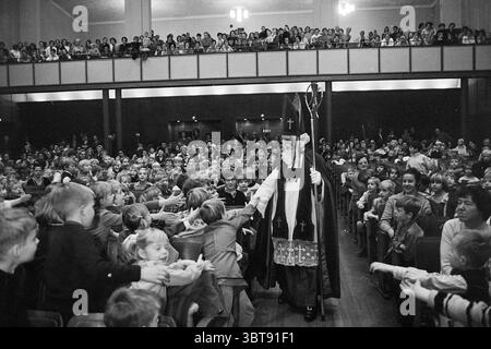 Sint Nicolaas Concertgebouw préparé Nicholas Sinterklaas, Whizgle News, Dutch Desk, The Netherlands, 1950 - 2000 on 04-12-1968. Ces rubriques apparaissent dans l'image. Dans un grand auditorium rempli d’un public, la scène est vivante avec énergie et anticipation. L'espace est bondé, avec des rangées de personnes s'étendant du premier plan vers l'arrière, soulignant l'ampleur du rassemblement. Au centre, un homme âgé, vêtu de vêtements ecclésiastiques traditionnels richement détaillés, se distingue. Sa tenue combine des robes sombres et fluides avec des embellissements ornés qui font allusion à son autorité et au sig Banque D'Images