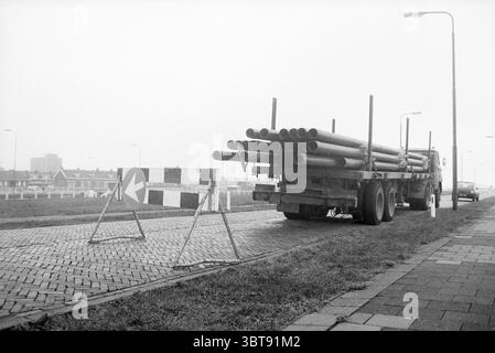 Camion avec tuyaux garés avec barrière d'avertissement, Whizgle News, Dutch Desk, pays-Bas, 1950 - 2000 en 1986. L'image inclut ces rubriques. La scène représente un environnement gris et brumeux, où la visibilité semble limitée. Un gros camion est garé bien en vue sur le côté gauche, chargé de nombreux longs tuyaux cylindriques empilés horizontalement. La surface métallique du chariot réfléchit des nuances de lumière atténuées en raison de l'environnement couvert. Devant le chariot se trouvent deux barrières de construction, peintes avec des bandes noires et blanches alternées, indiquant une zone restreinte. Le bar Banque D'Images