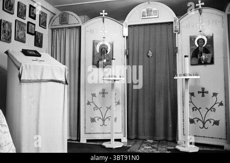 Église orthodoxe russe dans la nef sur la Spaarne Haarlem pays-Bas, Whizgle News, Dutch Desk, pays-Bas, 1950 - 2000 le 14-08-1962. L'image montre ces rubriques. La scène dépeint un espace intérieur serein et intime, probablement une petite chapelle ou un espace dédié au culte. Les murs sont ornés de grandes icônes encadrées qui représentent diverses figures religieuses, offrant une riche tapisserie de couleurs et de détails complexes. Au premier plan, un autel simple est visible, drapé d'un tissu doux et texturé, probablement blanc ou clair, soulignant la pureté et la révérence. L'autel est fixé Banque D'Images