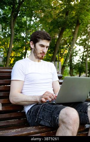 Jeune homme en t-shirt blanc travaillant sur ordinateur portable dans le parc. Vue latérale d'un gars tapant sur le banc à l'extérieur. Banque D'Images
