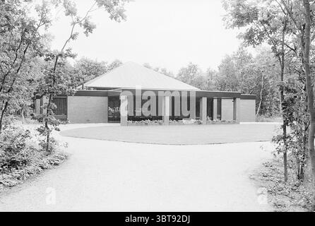 Extérieur d'un auditorium dans un cimetière/crématorium., Whizgle News, Dutch Desk, pays-Bas, 1950 - 2000. L'image contient ces rubriques. La scène présente une structure architecturale moderne située dans un paysage verdoyant et luxuriant. Le bâtiment dispose d'un toit plat et angulaire qui s'étend vers l'extérieur, lui donnant une silhouette saisissante contre le ciel. Ses murs sont composés de briques rouges et de grands panneaux de verre, permettant à la lumière naturelle de pénétrer l'intérieur tout en offrant une vue sur les environs. Devant le bâtiment, une pelouse soigneusement entretenue s’étend, bordée d’un jardin à plan fleuri Banque D'Images