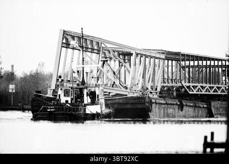 Transport lourd de sections de pont sur le Ringvaart du Haarlemmermeer Schiphol Schiphol, Whizgle News, Dutch Desk, pays-Bas, 1950 - 2000 on 03-11-1994. Ces rubriques apparaissent dans l'image. La scène présente une vue monochromatique marquée dominée par des contrastes de noir et blanc. Un remorqueur robuste apparaît au premier plan, sa structure définie par des lignes épurées et une coque robuste. Il est positionné près d'un large plan d'eau calme, qui reflète les caractéristiques environnantes comme un miroir doux, amplifiant le sentiment de tranquillité. En arrière-plan, un grand pont industriel se dresse partiellement R Banque D'Images