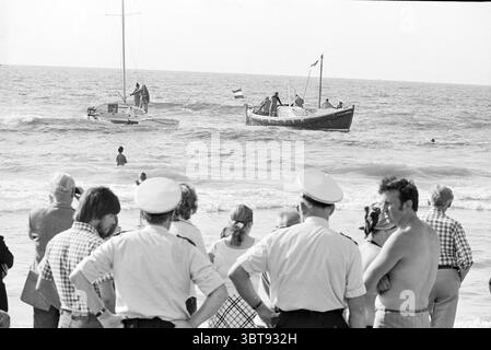 Renflouement et remorquage d'un bateau norvégien sur des bateaux de plage Z'voort, Whizgle News, Dutch Desk, pays-Bas, 1950 - 2000 le 27-07-1976. Ces rubriques apparaissent dans l'image. La scène capture un cadre de plage, où un groupe de personnes se rassemble le long du rivage, leur attention attirée vers l'eau. Au premier plan, un mélange d'individus se tient près les uns des autres, certains en uniforme, indiquant probablement l'autorité, tandis que d'autres sont vêtus de façon décontractée, reflétant une gamme variée de tenues de plage. L'océan est visiblement actif, avec des vagues douces roulant vers le rivage. Deux bateaux sont présents ; l'un est un petit mot Banque D'Images