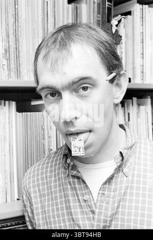 Joost Swarte Cartoonist dessins, Whizgle News, Dutch Desk, pays-Bas, 1950 - 2000 le 14-08-1985. L'image montre ces rubriques. Dans la scène, un homme se tient avec une expression quelque peu ludique, mettant en valeur un détail unique : un petit objet rectangulaire caché dans sa bouche. Sa coiffure semble légèrement bouleversée, ajoutant à un comportement décontracté. Sa chemise, avec un motif à carreaux dans les tons de bleu et de blanc, contraste avec la toile de fond, qui est remplie de rangées de livres et de disques, suggérant une bibliothèque ou une collection musicale bien garnie et éventuellement personnelle. L'éclairage est doux, par Banque D'Images