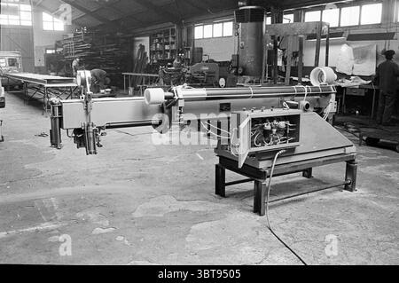 Factory Hall., Whizgle News, Dutch Desk, pays-Bas, 1950 - 2000. L'image montre ces rubriques. La scène se déroule dans un atelier spacieux caractérisé par un haut plafond voûté. La lumière naturelle pénètre à travers de grandes fenêtres, projetant des ombres douces sur le sol en béton, ce qui révèle un mélange de poussière et d'usure résultant de plusieurs années d'utilisation. Au premier plan, une grande machine industrielle se tient en évidence. Son cadre robuste est peint en gris discret, accentué par des composants métalliques usés qui font allusion à une histoire de dur labeur. La machine est équipée de différents leviers et tubes, suggérant sa fonctio complexe Banque D'Images