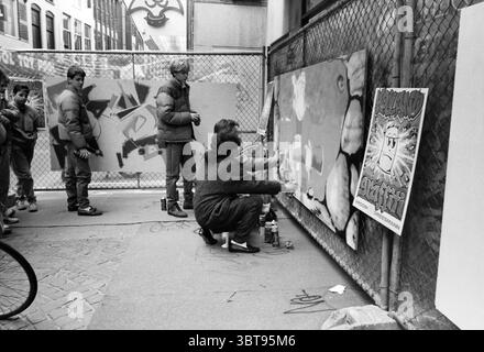 Graffiti chez V&D. Dessins dessins, Whizgle News, Dutch Desk, pays-Bas, 1950 - 2000 le 02-04-1987. L'image montre ces rubriques. En milieu urbain, un groupe d'individus est rassemblé dans une ruelle étroite, flanquée de grands bâtiments qui jettent un sentiment d'enfermement. La scène capture un moment d'expression artistique, où les couleurs vibrantes de l'art graffiti sont créées sur de grandes toiles fixées contre une clôture à maillons de chaîne. Une femme, accroupie, peint activement, sa figure concentrée et engagée. Elle porte un pull sombre et surdimensionné qui contraste avec ses boîtes de peinture aux couleurs vives n Banque D'Images