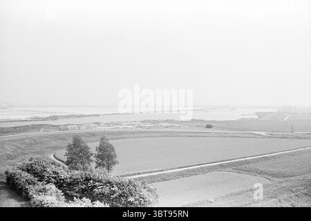 Travaux d'élargissement du canal de la mer du Nord ports Ouest Amsterdam North Holland villages etc. North Sea canal Works, Whizgle News, Dutch Desk, pays-Bas, 1950 - 2000 on 26-05-1964. L'image inclut ces rubriques. La scène présente un vaste paysage tranquille avec des tons discrets de noir et blanc, créant une atmosphère douce et rêveuse. Au premier plan, des champs roulants d'herbe et de cultures s'étendent à travers la terre, affichés dans différentes nuances de gris. Une courbe douce dans le chemin de terre suggère un voyage tranquille, conduisant le regard à travers l'étendue tranquille. À gauche, un amas de buissons bas fournit un naturel Banque D'Images