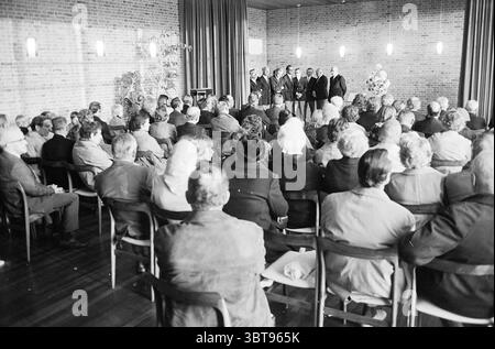 Journée du souvenir, Whizgle News, Dutch Desk, pays-Bas, 1950 - 2000 en 1973. Ces rubriques sont illustrées dans l'image. La scène représente un rassemblement dans une pièce spacieuse et bien éclairée caractérisée par des tons chauds provenant des murs de briques et du parquet. Un groupe de six individus se tient à l'avant, formant un demi-cercle autour d'un arrangement floral, qui ajoute une touche de couleur et de vitalité à la palette autrement neutre. Ces personnes semblent être engagées dans un moment solennel ou cérémoniel, peut-être prononcer un discours ou honorer quelqu'un. Le public, composé à la fois d'hommes et de femmes, est s. Banque D'Images