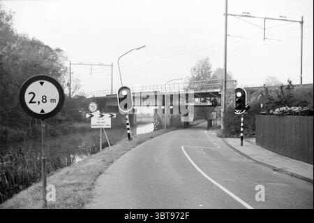 Pont ferroviaire V'zang Vogelenzang, Whizgle News, Dutch Desk, pays-Bas, 1950 - 2000 on 16-10-1996. Ce sont les éléments de l'image. La scène présente un paysage urbain tranquille avec un accent sur un pont enjambant une étendue d'eau calme. Le pont est construit en métal et semble quelque peu industriel, présentant une texture altérée. Sa structure est soutenue par des piliers en béton, et une végétation verte est visible autour des bords, laissant entendre son intégration avec la nature. Au premier plan, une route sinueuse mène vers le pont. Le chemin est bordé par une clôture en bois sombre Banque D'Images