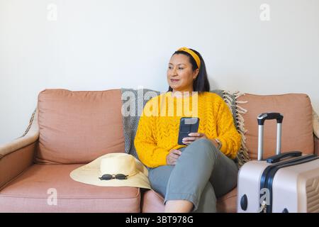 Femme portant un pull jaune assis sur le canapé droit à la maison tenant le téléphone près de la valise, espace de copie Banque D'Images
