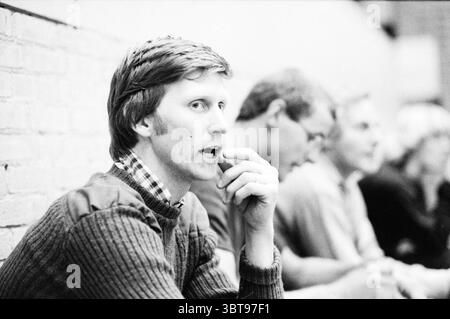 Portrait entraîneur de football en salle partenaire du maillot féminin : Ten Cate BV distribution Agency Alkmaar, Whizgle News, Dutch Desk, pays-Bas, 1950 - 2000 on 10-1980. Voici les rubriques de l'image. Dans un intérieur légèrement éclairé, un homme est assis à une table, regardant soigneusement au loin. Ses cheveux sont clairs et soignés, jumelés à un pull texturé et sombre qui contraste avec les tons doux autour de lui. Il tient une main à sa bouche, suggérant une profonde contemplation ou peut-être une hésitation. Derrière lui, plusieurs autres individus sont visibles, légèrement floues pour indiquer qu'ils font partie d'une sce plus grande Banque D'Images