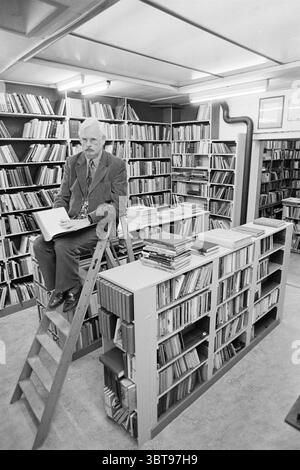 Mr. Hovingh dans sa librairie d'antiquaires Haarlem Kleine Houtstraat 50 pays-Bas, Whizgle News, Dutch Desk, pays-Bas, 1950 - 2000 le 12-11-1995. Ces rubriques sont illustrées dans l'image. La scène présente un environnement de bibliothèque confortable mais organisé avec de grandes étagères remplies d'une myriade de livres, créant une toile de fond texturée de couleurs et de motifs variés. Les étagères dominent l'espace, avec des livres empilés étroitement ensemble, certains debout tandis que d'autres sont à plat. Les teintes prédominantes sont les bruns et les verts chauds, contribuant à une atmosphère savante. Au premier plan, un pers Banque D'Images