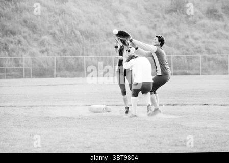 Pino's - Spaarnwoude Softball, Whizgle News, Dutch Desk, pays-Bas, 1950 - 2000 le 18-08-1979. Voici les rubriques de l'image. La scène capture un moment énergique sur un terrain de sport, où trois joueurs sont engagés dans un jeu dynamique. Au premier plan, un athlète, portant un maillot blanc, est pris en pleine action, tentant de marquer un autre joueur tout en s'accroupissant au sol. Le deuxième joueur, vêtu d'un maillot sombre, saute avec une expression déterminée, les mains atteignant une balle molle qui est lancée au-dessus de lui. Le troisième individu, positionné légèrement derrière, montre un mix Banque D'Images