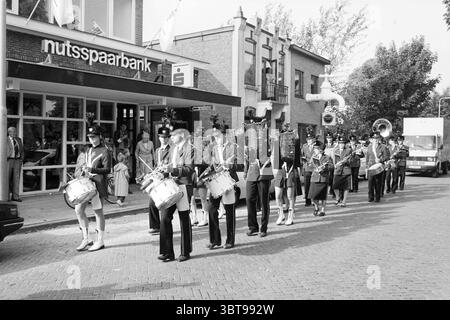 Journée portes ouvertes Nutsspaarbank Bennebroek Nuts United Savings Banks Bennebroek, Whizgle News, Dutch Desk, pays-Bas, 1950 - 2000 on 05-10-1985. L'image montre ces rubriques. La scène présente un défilé marchant dans une rue large, remplie d'une atmosphère animée. Les marcheurs sont vêtus d'uniformes de style militaire, avec un mélange de couleurs sombres et claires, principalement des nuances de gris et de noir. Certains individus portent des instruments, affichant bien en évidence des tambours et un tuba, tandis que d'autres marchent de manière synchronisée, maintenant une formation précise. D'un côté de la rue, un contemporain Banque D'Images
