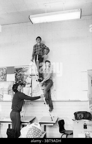 Pigeons dans la lampe dans la salle de classe Dreefschool Wilhelminastraat Vogels Haarlem Wilhelminastraat pays-Bas, Whizgle News, Dutch Desk, pays-Bas, 1950 - 2000 le 24-06-1982. L'image contient ces rubriques. Dans la scène, deux garçons sont positionnés sur une échelle blanche, qui est appuyée contre un mur affichant une variété de cartes et d'affiches. Un garçon se tient confiant en haut, portant une chemise à carreaux, tandis que l'autre, plus bas, regarde vers le haut, vêtu de façon décontractée. Les garçons montrent un sentiment de camaraderie et de détermination alors qu'ils s'engagent dans une tâche impliquant les décorations murales. Une femme se tient à t Banque D'Images