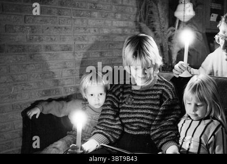 Panne de courant à Haarlem North Lighting Haarlem pays-Bas, Whizgle News, Dutch Desk, pays-Bas, 1950 - 2000 le 20-11-1984. Ces rubriques sont illustrées dans l'image. Dans un intérieur confortable, une famille est réunie, créant une scène remplie de chaleur et d'intimité. Le cadre présente un mur de briques texturées, qui sert de toile de fond, ajoutant un charme rustique à l'atmosphère. Un éclairage doux émane de deux bougies tenues par des adultes, projetant une douce lueur qui met en valeur les visages des enfants et de la femme au centre. La femme, portant un pull tricoté avec un motif distinct, respire Banque D'Images
