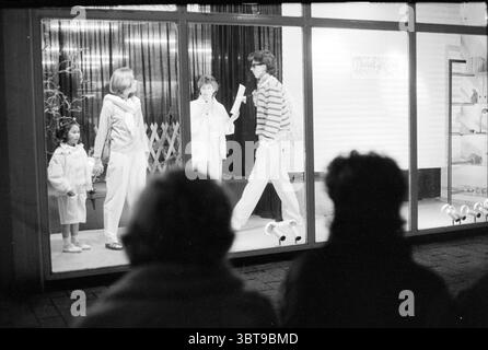 Défilé de mode dans la fenêtre Anegang Fashion Haarlem Anegang The Netherlands, Whizgle News, Dutch Desk, The Netherlands, 1950 - 2000 le 28-03-1985. Ce sont les éléments de l'image. La scène se déroule dans une zone urbaine animée, présentant une vitrine qui capte l'attention des passants. À l'intérieur du boîtier en verre éclairé, trois figurines sont bien en évidence. Deux adultes et un enfant, tous vêtus d'une tenue élégante mais décontractée, font preuve d'un sentiment de jeu et d'engagement. L'enfant, debout à gauche, enrobe une tenue de couleur claire, avec un design ludique, qui contraste doucement Banque D'Images
