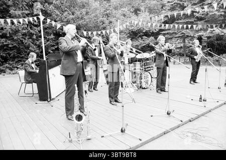 Dutch swing College band open air Theatre Music, Whizgle News, Dutch Desk, pays-Bas, 1950 - 2000 le 04-07-1986. L'image montre ces rubriques. La scène se déroule sur une scène en bois, entourée d'une végétation luxuriante qui suggère un cadre extérieur vibrant. Un groupe de six musiciens, vêtus de costumes noirs formels, se positionnent en ligne, profondément engagés dans leur performance. Chaque musicien joue d'un instrument de cuivres différent, contribuant à une atmosphère animée. Au premier rang se trouvent un musicien avec un saxophone, un autre jouant de la trompette, tandis qu'un troisième se tient derrière eux, prêt au micropho Banque D'Images