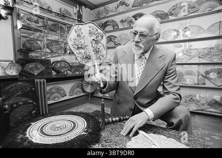 Mr. Tax avec collection de fans fans, Whizgle News, Dutch Desk, pays-Bas, 1950 - 2000 sur 21-05-1974. L'image inclut ces rubriques. Dans une pièce chaleureusement éclairée remplie d'un éventail de ventilateurs décoratifs tapissant les murs, un homme âgé est assis à une table en bois. Sa tenue se compose d'un costume de couleur claire complété par une cravate à motifs, dégageant un sentiment d'élégance et de sophistication. Ses cheveux blancs contrastent avec les nuances plus sombres de ses vêtements, et ses expressions faciales véhiculent un sentiment de contemplation et de nostalgie. Dans sa main droite, il tient un ventilateur de main finement conçu, orné d'esprit Banque D'Images