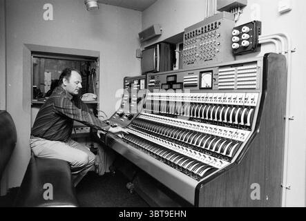 M.R. Faassen dans le stand Schouwburg Haarlem Schouwburg Haarlem Wilsonsplein pays-Bas, Whizgle News, Dutch Desk, pays-Bas, 1950 - 2000 le 09-08-1978. Ce sont les éléments de l'image. Dans une pièce remplie de tons discrets et d'une atmosphère nostalgique, un homme est assis concentré sur un grand panneau de commande au design complexe qui domine le premier plan. Le panneau comporte de nombreux boutons et curseurs, disposés méthodiquement, avec un mélange de textures métalliques et plastiques qui évoquent un sens de la technologie vintage. L'homme, vêtu d'une chemise à carreaux, se penche légèrement en avant, sa main droite adj Banque D'Images