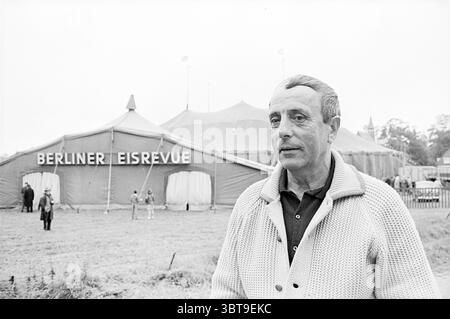 Ernst Bayer Persons, Whizgle News, Dutch Desk, pays-Bas, 1950 - 2000 le 12-07-1964. Ces rubriques sont illustrées dans l'image. La scène montre un homme debout bien en vue au premier plan, portant un cardigan de couleur claire sur une chemise sombre, qui contraste avec les tons doux de l'arrière-plan. Son expression est calme mais légèrement contemplative, avec une lumière douce illuminant ses traits, soulignant les lignes douces de son visage. Derrière lui, une grande tente domine le fond, sa structure soulignée par une teinte riche et vibrante, éventuellement un rouge ou un orange chaud. La conception de la tente est cla Banque D'Images