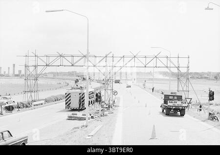 Échafaudage pour Velsertunnel Signs Works, Whizgle News, Dutch Desk, pays-Bas, 1950 - 2000 le 01-07-1965. Ces rubriques apparaissent dans l'image. La scène capture une large chaussée coupant en deux une étendue ouverte, caractérisée par une palette de niveaux de gris qui souligne le contraste. Au premier plan, une grande structure d'échafaudage partiellement construite s'élève au-dessus de la route, laissant entendre la présence de travaux en cours ou d'un événement potentiel. L'échafaudage est composé de poutres métalliques, formant une arche qui suggère un point d'entrée cérémoniel ou point de contrôle. D'un côté de la route, un véhicule de taille, peut-être un camion Banque D'Images