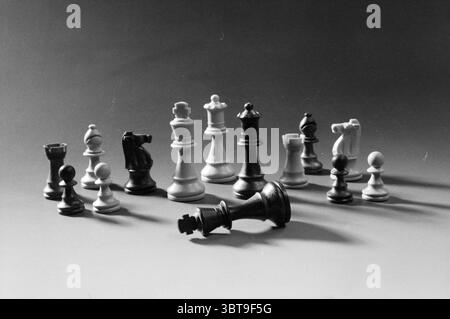 Chess PIECES., Whizgle News, Dutch Desk, pays-Bas, 1950 - 2000. Ces rubriques apparaissent dans l'image. La scène présente une collection de pièces d'échecs disposées sur une surface lisse, éclairées par un éclairage doux et diffus qui projette des ombres douces, améliorant l'effet tridimensionnel. La disposition est asymétrique, avec plusieurs pièces debout fièrement debout tandis qu'un roi éminent s'est renversé, attirant l'attention sur sa chute. Les pièces d'échecs sont un mélange de noir et blanc, contrastant fortement avec le fond neutre, qui passe des tons clairs aux tons sombres, ajoutant de la profondeur et du drame. Th Banque D'Images