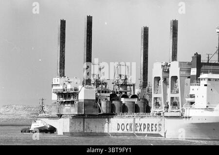 Boot Dock Express Approaches IJmuiden Locks IJmuiden pays-Bas, Whizgle News, Dutch Desk, pays-Bas, 1950 - 2000 on 10-04-1985. Ces rubriques apparaissent dans l'image. La scène capture un grand navire caractérisé par ses caractéristiques industrielles. Dominant le premier plan, le navire a une coque lourde et altérée, peinte dans des tons de gris qui se fondent avec le ciel silencieux. Plusieurs grues imposantes, très proéminentes contre l'horizon, se dressent au sommet du navire, leurs structures métalliques créant un sentiment de verticalité. Au premier plan, un petit remorqueur est positionné à côté du plus grand navire, IT Banque D'Images