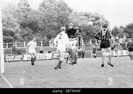 Football Noorder Sportpark Haarlem pays-Bas., Whizgle News, Dutch Desk, pays-Bas, 1950 - 2000. L'image montre ces rubriques. La scène capture un moment intense sur un terrain herbeux pendant un match de football. Un groupe de joueurs est engagé dans l'action, avec un joueur, vêtu d'un maillot sombre, sautant pour intercepter une balle juste au-dessus de la ligne de but. Son expression est concentrée et déterminée, laissant entendre la pression du moment. Autour de lui, deux joueurs en maillots blancs et rayés contrastés sont positionnés stratégiquement. Le joueur portant le maillot numéro 2 semble surveiller le Banque D'Images
