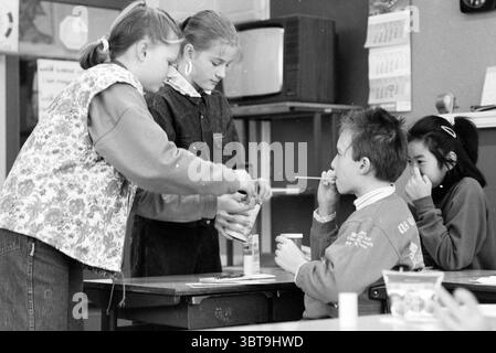 Discours CARA Eenhoornschool, Whizgle News, Dutch Desk, pays-Bas, 1950 - 2000 on 10-02-1991. L'image montre ces rubriques. Dans une salle de classe, quatre enfants participent à une activité collaborative. L'atmosphère est animée et remplie d'un sentiment de curiosité et de jeu. Deux filles se tiennent près d'un bureau, l'une portant un pull bleu vif orné d'un motif floral, tandis que l'autre est dans une tenue noire classique, les deux affichant des expressions ciblées en interagissant étroitement. La fille dans le pull floral verse le contenu d'un récipient dans une petite tasse, son attention clairement Banque D'Images