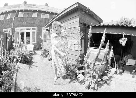 Garden Pride of MRS Nulte Haarlem pays-Bas, Whizgle News, Dutch Desk, pays-Bas, 1950 - 2000 on 10-06-1992. L'image montre ces rubriques. Dans un jardin ensoleillé, une femme âgée se tient bien en vue, serrant un râteau. Elle porte une robe à pois, qui ajoute une touche de nostalgie à la scène. Ses cheveux sont gris, coiffés d'une manière douce et bouclée, reflétant la douce lumière du jour. Autour d'elle se trouvent diverses plantes et fleurs, principalement dans des verts vibrants, avec certaines fleurs dans des couleurs douces qui améliorent l'atmosphère accueillante du jardin. En arrière-plan, un hangar en bois complète t Banque D'Images