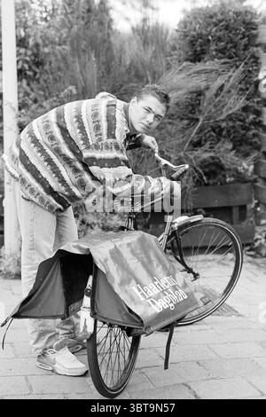 Livreur de journaux le quotidien de Haarlem, Whizgle News, Dutch Desk, pays-Bas, 1950 - 2000 en 1995. Ce sont les éléments de l'image. Dans un cadre extérieur animé, un jeune homme se penche sur un vélo, ajustant ses composants avec une expression ciblée. Il porte un pull coloré à motifs qui présente un mélange vibrant de rayures et de tourbillons, mettant en valeur un mélange de couleurs vives contre les tons plus discrets de son Jean denim. Le vélo a un sac de chargement distinctif attaché à elle, suggérant son utilisation pour le transport d'articles. Le fond est rempli de verdure luxuriante, avec bien entretenu Banque D'Images