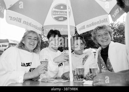 Band set Pint Bloemendaal Bloemendaal, Whizgle News, Dutch Desk, pays-Bas, 1950 - 2000 le 26-05-1990. Ces rubriques apparaissent dans l'image. Une scène animée en plein air met en scène quatre jeunes adultes réunis autour d'une table en bois à l'ombre d'un grand parapluie orné d'un logo. Le groupe se compose de deux femmes et deux hommes, tous affichant des expressions joyeuses, soulignant un sentiment de camaraderie. La première femme, à gauche, a les cheveux courts et blonds clairs et porte un haut blanc avec un léger rouleau au col. Son sourire éclatant rayonne de chaleur et de convivialité. A côté d'elle se trouve un homme avec Banque D'Images