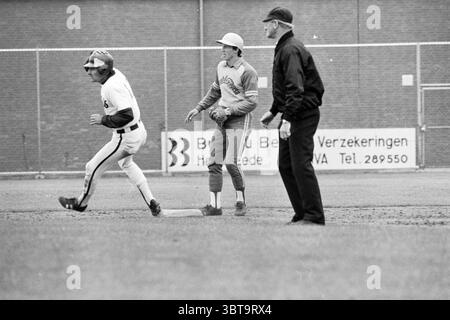 Nicols - Amstel Tigers Baseball Haarlem Nicols, Whizgle News, Dutch Desk, pays-Bas, 1950 - 2000 le 24-05-1980. L'image contient ces rubriques. La scène représente un match de baseball dans un cadre animé et extérieur. Au premier plan, un joueur portant un uniforme blanc avec des rayures noires court vers la deuxième base. Son expression transmet la détermination, et sa casquette est légèrement inclinée, suggérant un éclat d'énergie pendant qu'il court. Le gazon sous lui est bien entretenu, avec une riche couleur verte contrastant avec la saleté brune de la base. À la deuxième base, un autre joueur, vêtu d'un uniforme gris Ado Banque D'Images