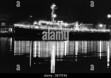 The Dashing One in the light, Whizgle News, Dutch Desk, pays-Bas, 1950 - 2000 on 13-12-1994. Ces rubriques sont illustrées dans l'image. La scène capture un front de mer tranquille la nuit, dominé par un grand navire qui semble être un navire ancré tranquillement. Le navire est orné d'une série de lumières scintillantes qui créent une lueur chaleureuse et invitante sur fond d'obscurité. Ces lumières se reflètent sur la surface de l'eau calme, générant un motif fascinant de lignes chatoyantes qui ondulent doucement avec le mouvement subtil de l'eau. Les environs se composent d'un mélange de structures le long Banque D'Images