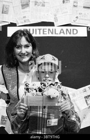 Cours dans le journal Hilversum, Whizgle News, Dutch Desk, pays-Bas, 1950 - 2000 sur 09-12-1994. L'image montre ces rubriques. Dans un cadre dynamique, une femme et un garçon se tiennent étroitement ensemble, dégageant un sentiment de chaleur et de camaraderie. La femme a les cheveux longs et foncés et porte une blouse de couleur claire jumelée à un gilet à motifs, créant un contraste saisissant avec le fond plus sombre. Son expression est lumineuse et accueillante, suggérant joie ou fierté. Le garçon à côté d'elle est plus jeune, portant une casquette de baseball ornée d'un logo et d'une chemise à carreaux qui ajoute de la texture à la scène. Il tient une empreinte Banque D'Images