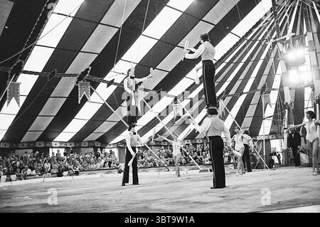 Toni Boltini dans Santpoort Circus, Whizgle News, Dutch Desk, pays-Bas, 1950 - 2000 sur 24-07-1974. Voici les rubriques de l'image. Dans une tente de cirque vibrante, l'atmosphère est remplie d'excitation et d'anticipation. Le tissu rayé du toit de la tente est un mélange de couleurs vives - rouges riches, bleus profonds et blancs vifs - créant un contraste dynamique avec l'intérieur faiblement éclairé. La lumière du soleil filtre à travers les ouvertures, projetant une lueur chaude sur la scène ci-dessous. Plusieurs artistes sont engagés dans un spectacle époustouflant d'acrobaties. L'un des interprètes s'équilibre précairement sur pilotis, dominant le Banque D'Images