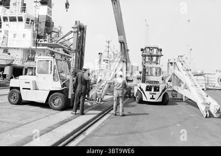 Engin de forage de chargement Velsen-Noord N.A.M. Velsen-Noord, Whizgle News, Dutch Desk, pays-Bas, 1950 - 2000 on 26-07-1983. Voici les rubriques de l'image. La scène représente une zone portuaire très fréquentée, remplie d'activité industrielle. Deux grandes machines, ressemblant à des chariots élévateurs ou à des manutentionnaires, dominent le premier plan. Ils sont peints dans un blanc dur et utilitaire, avec des marques noires proéminentes. Les véhicules ont une conception robuste et fonctionnelle, montrant des signes d'usure, ce qui indique leur utilisation fréquente dans des environnements difficiles. À proximité de la machinerie, deux travailleurs sont engagés Banque D'Images