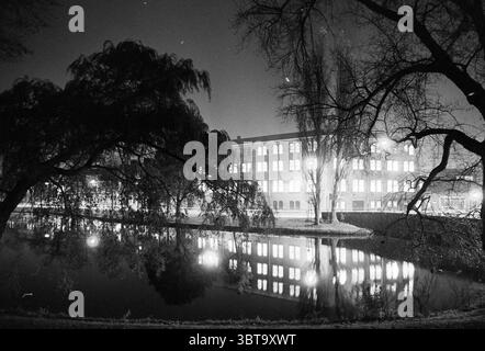 Kunstlijn 1993 : immeuble de bureaux illuminé Olympia Kinderhuissingel 6 Haarlem Kinderhuissingel pays-Bas, Whizgle News, Dutch Desk, pays-Bas, 1950 - 2000 on 02-11-1993. Ces rubriques sont illustrées dans l'image. La scène est dominée par une ambiance sereine et nocturne, caractérisée par une rivière qui reflète l'environnement avec une surface vitreuse. L'eau reflète la douce lueur des lumières d'un bâtiment en face, dont les grandes fenêtres émettent une teinte chaleureuse et invitante qui contraste magnifiquement avec l'obscurité de la nuit. Au premier plan, les branches de deux arbres s'étendent, leur silh Banque D'Images