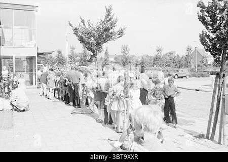 1st Customers Stuif Stuif Stuif Stuif Stuif, Whizgle News, Dutch Desk, pays-Bas, 1950 - 2000 le 14-07-1969. Voici les rubriques de l'image. La scène capture un cadre extérieur animé, caractérisé par une longue file de personnes qui attendent leur tour. À gauche, un groupe est rassemblé étroitement, avec quelques individus assis sur le sol, peut-être au repos tandis que d'autres se tiennent debout. Les personnes en ligne sont principalement des enfants et des adolescents, habillés dans une variété de styles vestimentaires qui reflètent une esthétique rétro. Leur tenue vestimentaire comprend des robes à motifs, des chemises rayées et des vestes légères aux couleurs pastel Banque D'Images