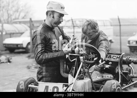 Courses de Kart sur une piste extérieure., Whizgle News, Dutch Desk, pays-Bas, 1950 - 2000. Ce sont les éléments de l'image. La scène capture deux individus engagés dans un moment de concentration et de collaboration. L'un, un homme adulte avec une moustache distinctive, est habillé d'un costume de course sombre, qui contraste avec les couleurs claires et délavées de l'environnement environnant. Son comportement suggère de l'expérience et des connaissances lorsqu'il examine un petit outil, probablement lié au kart sur lequel ils travaillent. La deuxième personne, un garçon plus jeune, se penche intensément sur le volant du Go-kart, vêtue d’une lumière Banque D'Images