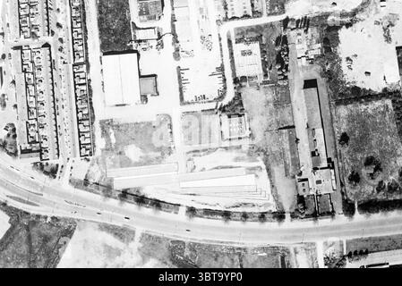 Récepteurs télescopiques avec homme, Whizgle News, Dutch Desk, pays-Bas, 1950 - 2000 en 1991. Ces rubriques sont illustrées dans l'image. La scène est une vue aérienne, mettant en valeur une zone développée avec quelques éléments naturels. D'un côté, il y a un long tronçon de route bordé par un mélange de bâtiments, y compris des unités résidentielles et des structures industrielles. La zone résidentielle se compose de nombreux petits bâtiments uniformes, probablement des maisons, disposés en rangées ordonnées, leurs toits formant un motif géométrique. À droite, un bâtiment industriel plus grand se distingue, avec un toit plat et un aménagement étendu. Banque D'Images